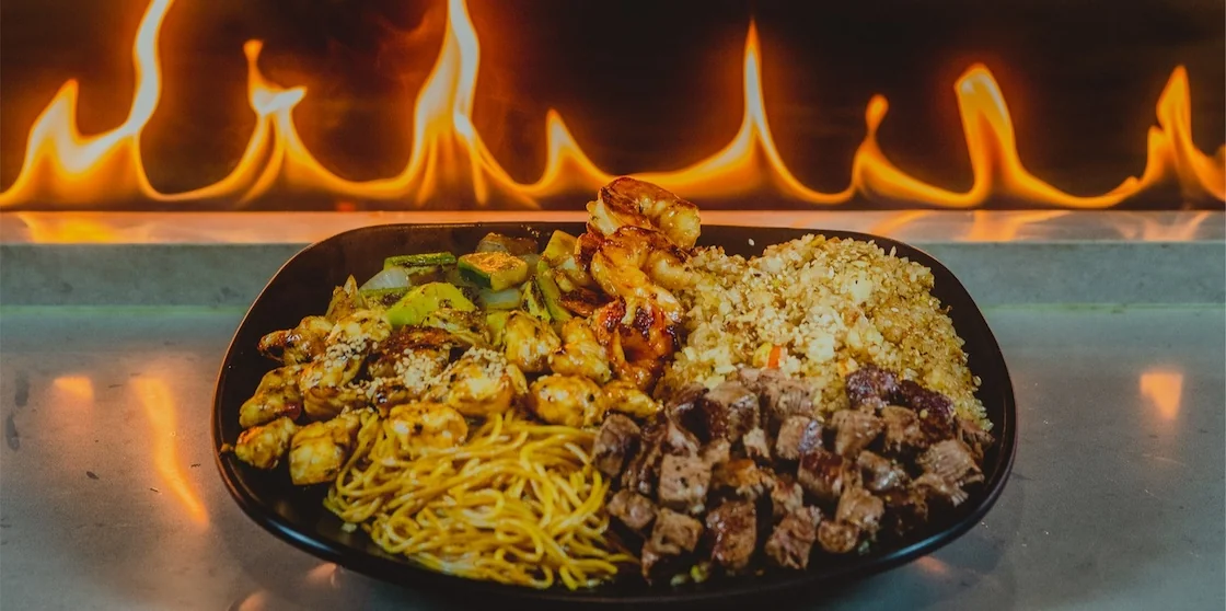 hibachi grill with flames behind a plate of noodles, rice, veggies, and meat