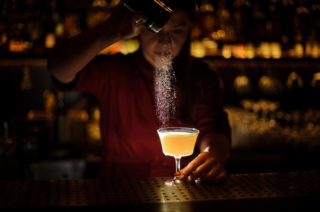 Female bartender serving a fresh delicious yellow cocktail and pouring a spice into it on the bar counter