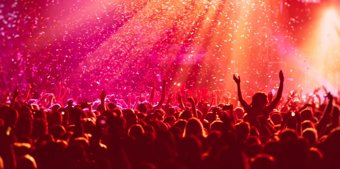 A crowded concert hall with scene stage in red lights, rock show performance, with people silhouette, colourful confetti explosion fired on dance floor during a concert festival - December concerts