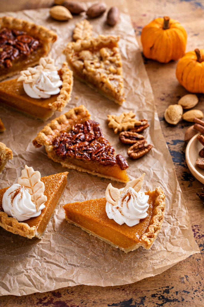 slices of pie - pumpkin, pecan, and more with pecans and pumpkins on a rough wooden table