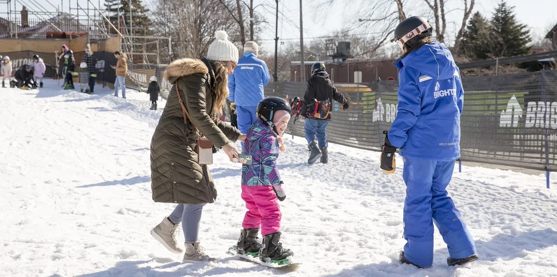 young girl learning how to snowboard with an instructor and parent along side her - february events