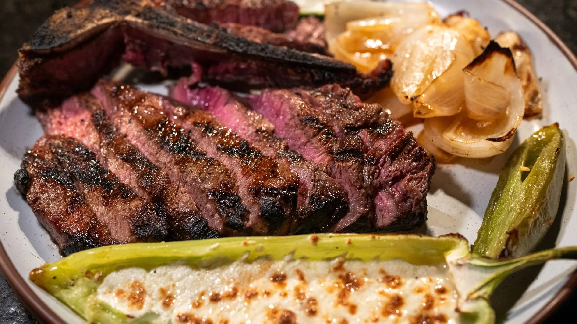 Top 10 - Vecino: alt="Sliced grilled steak cooked medium-rare on a ceramic plate, served with charred onions, roasted green vegetables, and a stuffed squash topped with melted cheese