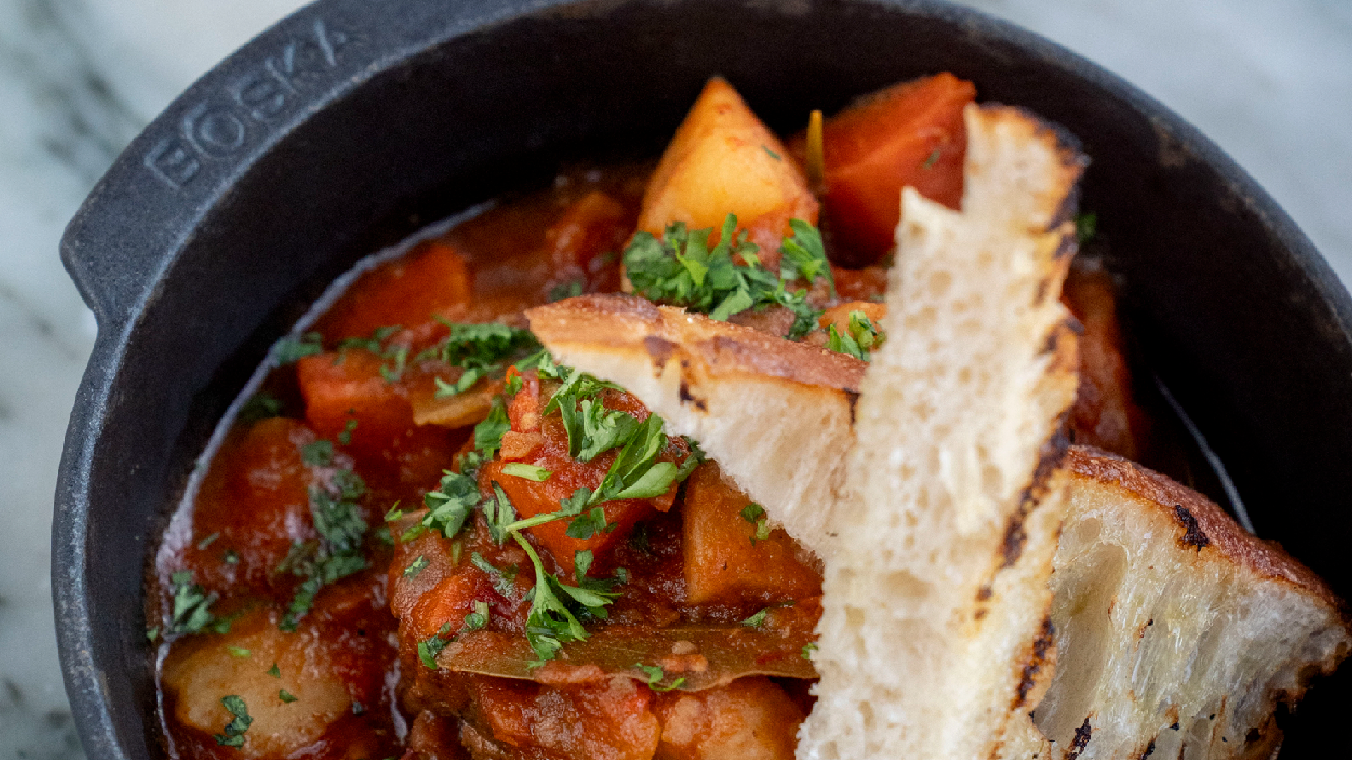Close-up of a cast-iron bowl filled with tomato-based stew containing vegetables, garnished with chopped herbs and served with toasted bread slices.