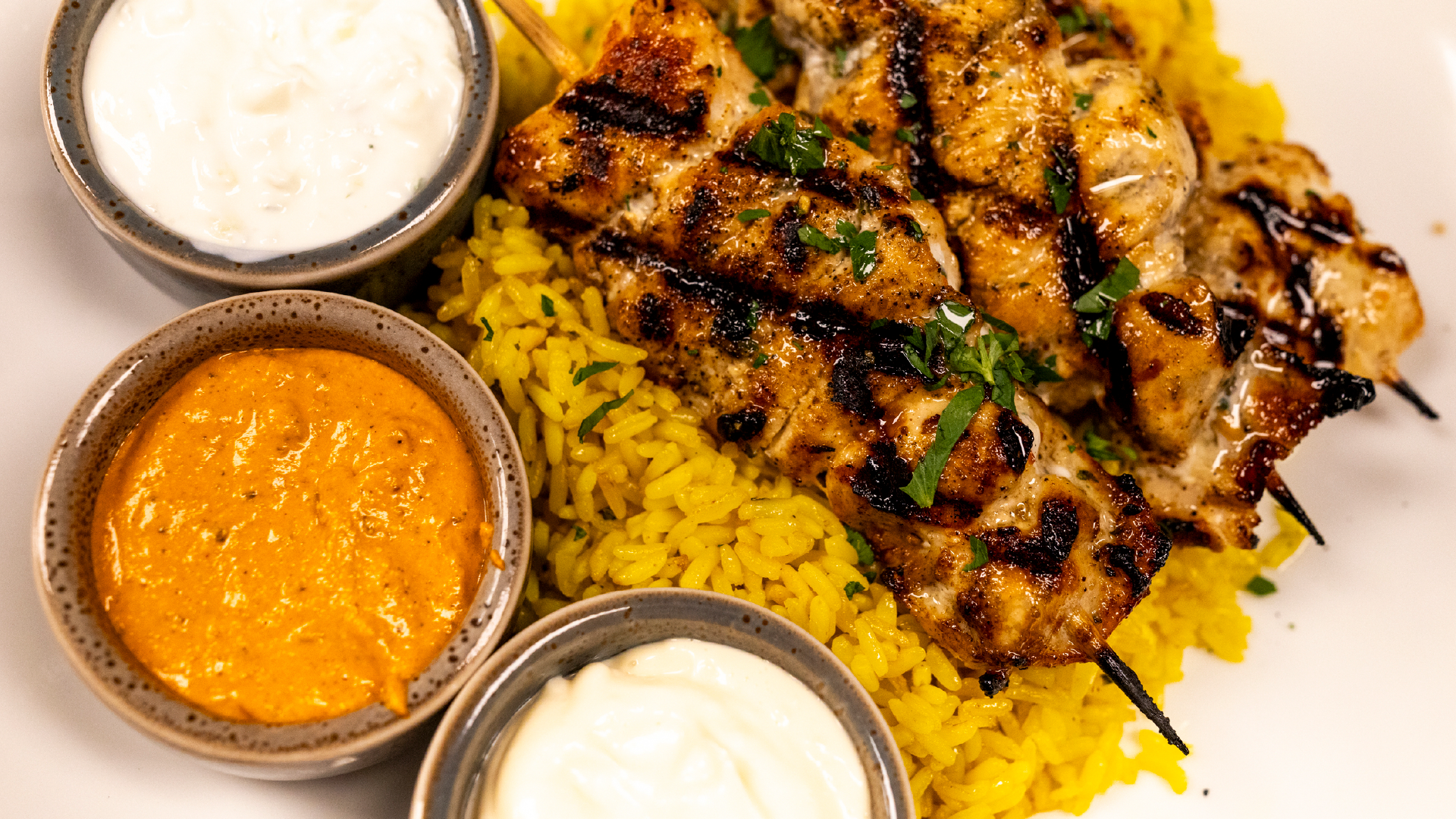 Grilled chicken skewers served over saffron rice with three dipping sauces including tzatziki and a spiced orange sauce, garnished with fresh herbs
