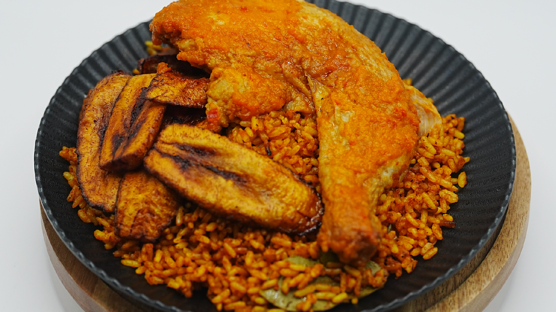 A generous serving of jollof rice topped with a whole seasoned chicken leg quarter and fried plantains, plated on a dark ridged dish set on a wooden coaster against a white background.