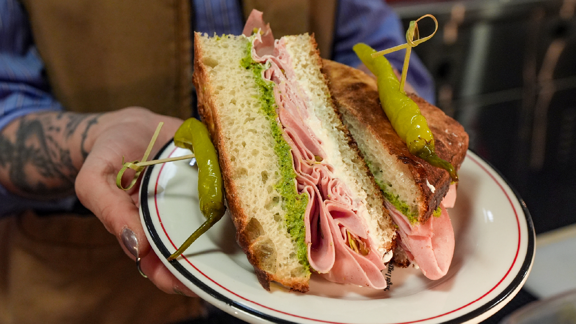 A tattooed server's hands holding a white diner plate with a ham sandwich on toasted sourdough bread, sliced in half and layered with green herb spread and thin-sliced pink ham, each half garnished with a pickled pepperoncini on a decorative toothpick