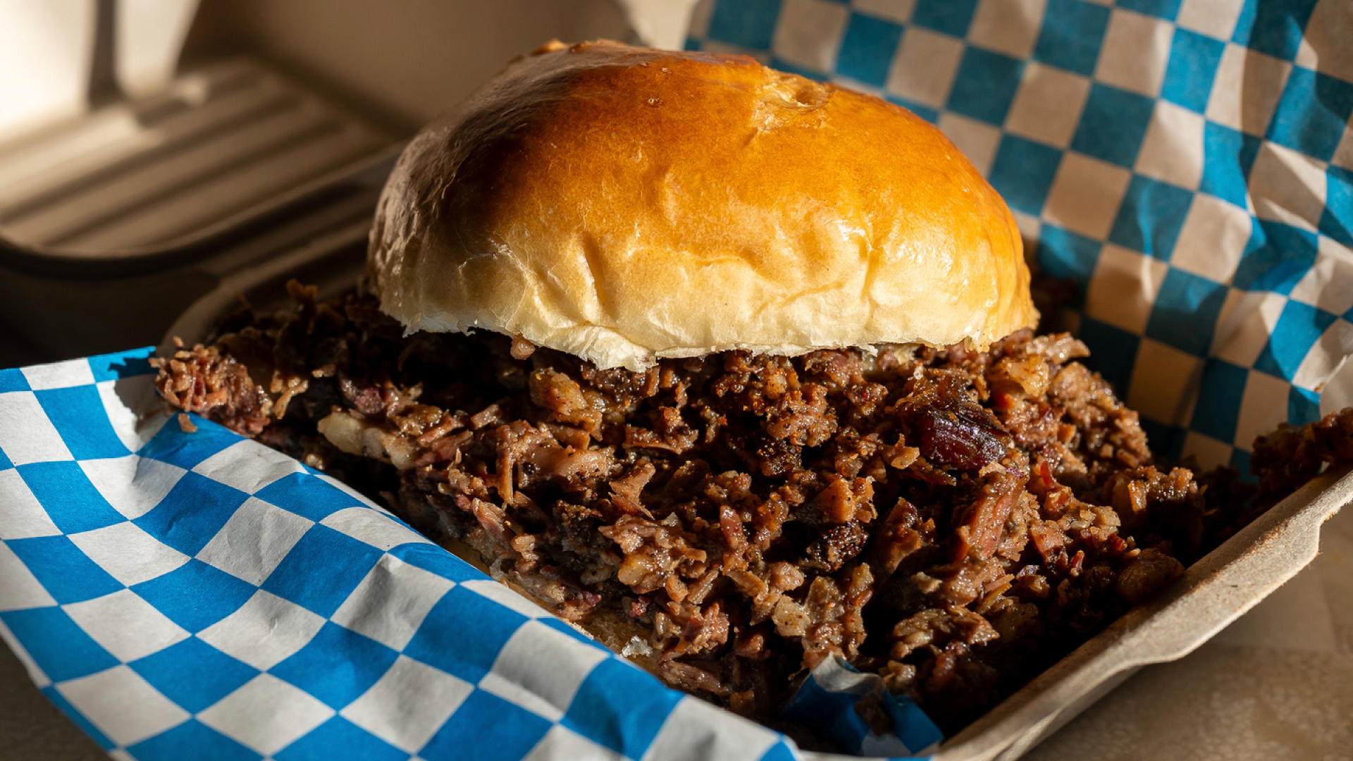 Close-up of a generously loaded pulled pork sandwich on a golden brioche bun, served in a blue-and-white checkered paper-lined basket at Blue Kuna Smokehouse.
