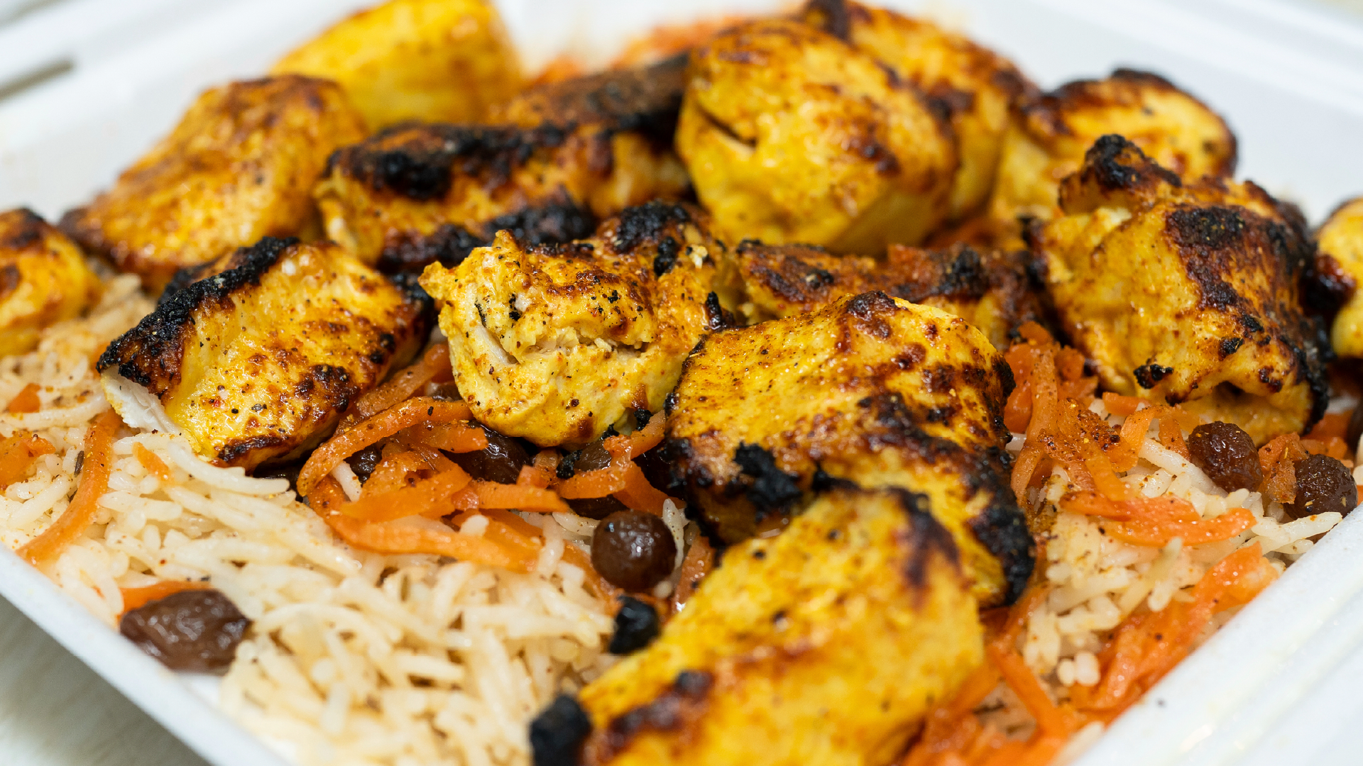 Close-up of a grilled chicken kebab dish from Ariana Afghan Restaurant, served over basmati rice with shredded carrots and raisins in a white takeout container.