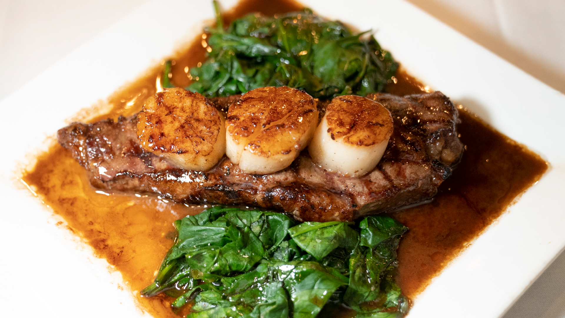 Close-up of a grilled steak topped with three golden seared scallops, served with sautéed spinach and a glossy brown pan sauce on a white square plate.