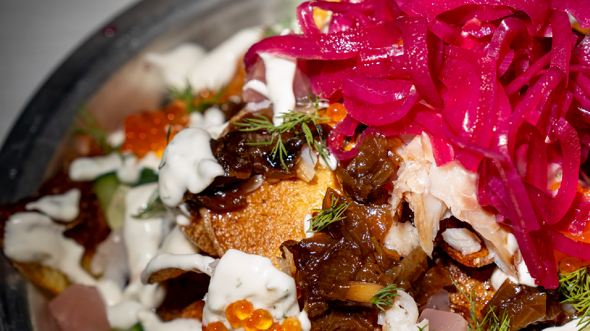 Close-up of a dish from Rose's Fine Food featuring crispy fritters, braised meat, salmon roe, herb crema, and shaved fennel, topped with vibrant pickled red onions.